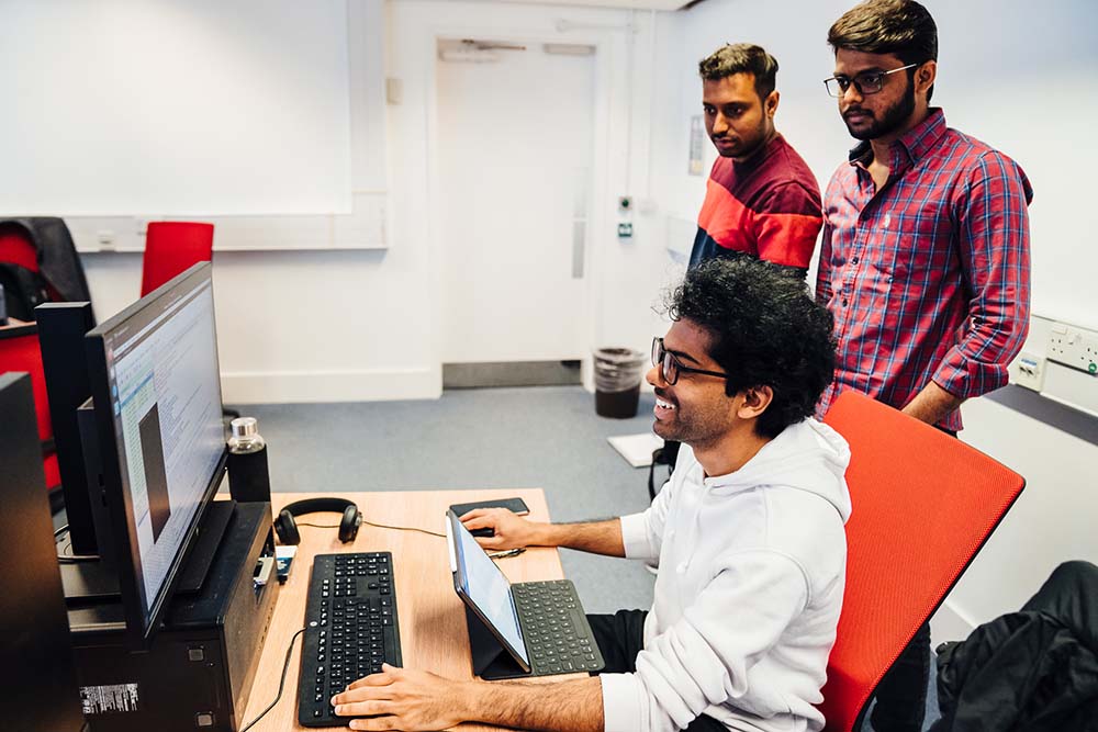 Cyber Security students in computer lab looking together at screen. One student is sat at desk with two others stood looking over his shoulder.