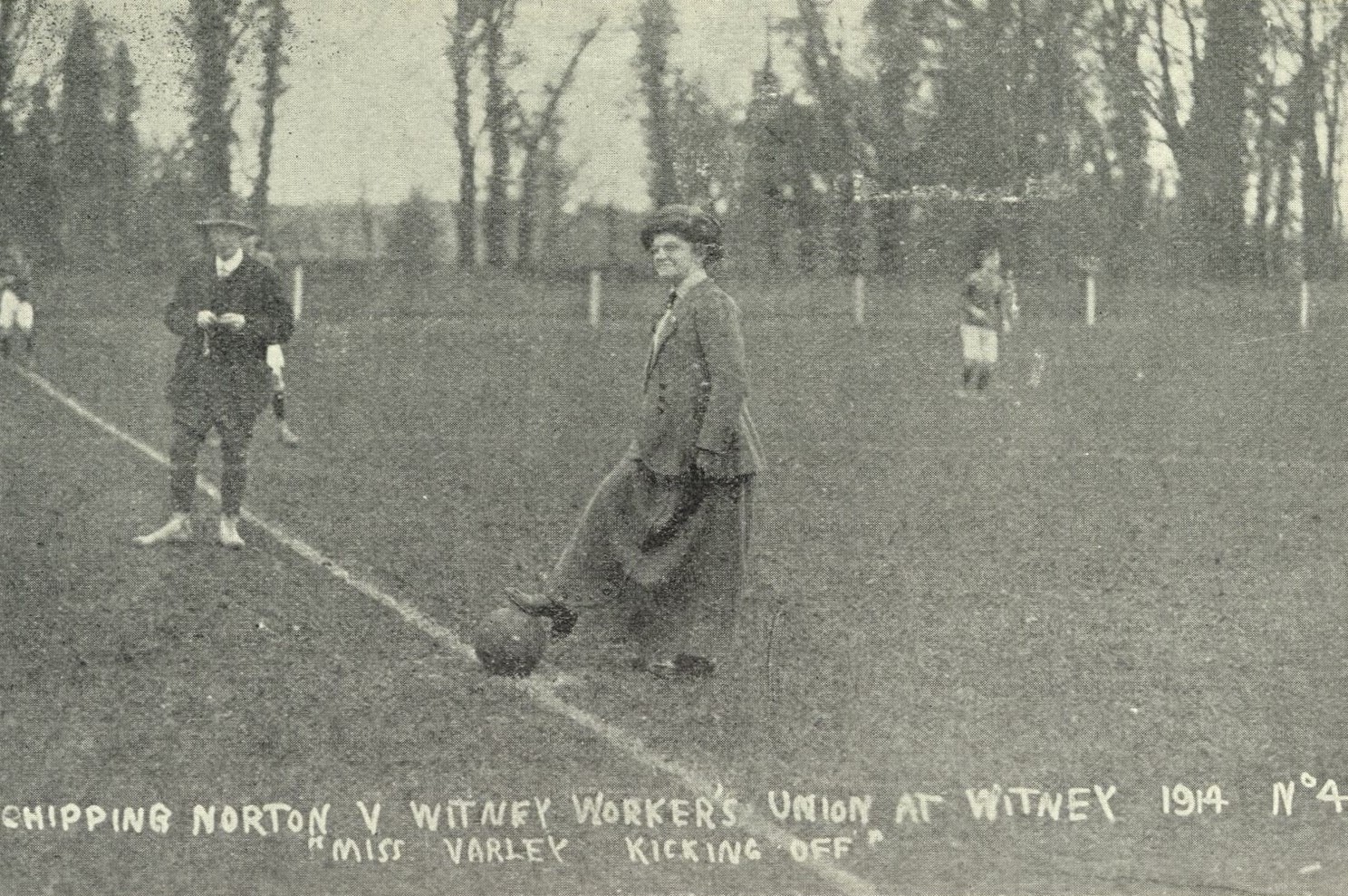Black and white photograph of Miss Varley kicking off the football match. She is standing in the middle of the football pitch with a foot on theball.
