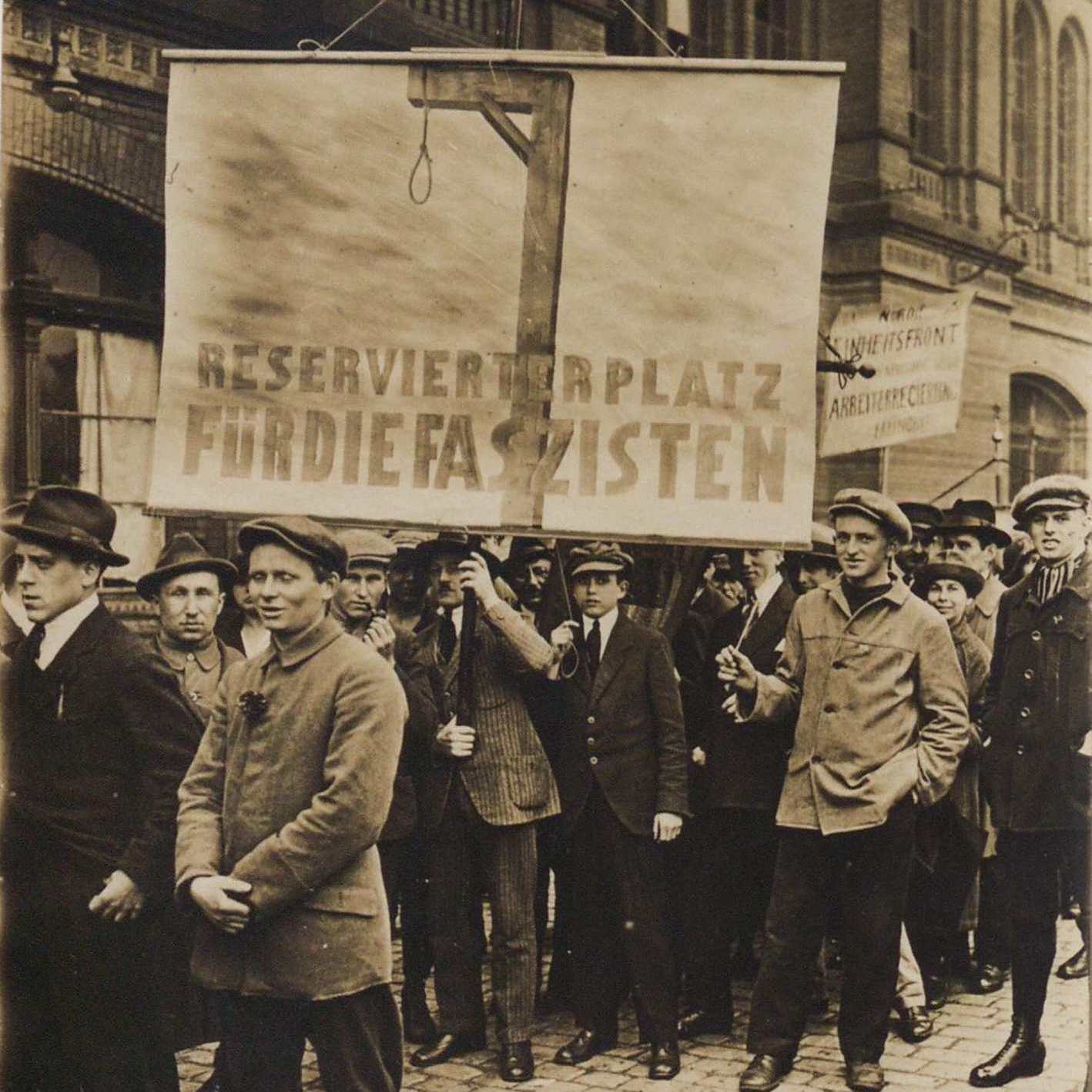 Group of men gathered on the street holding up a banner that portrays a gallows and the writing "RESERVIERTERPLATZ FURDIEFASZISTEN"