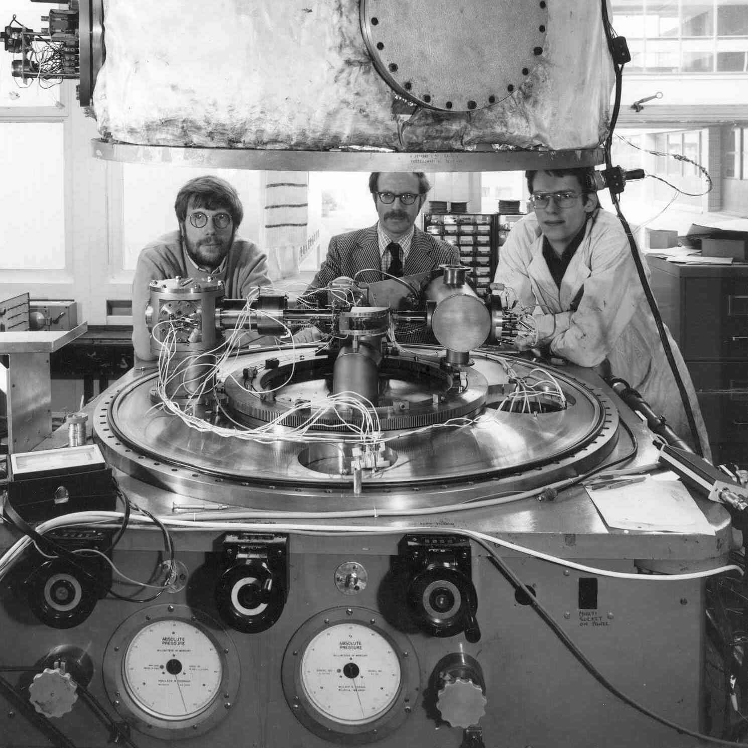 Black and white photograph of three men standing by a large machine in a science laboratory