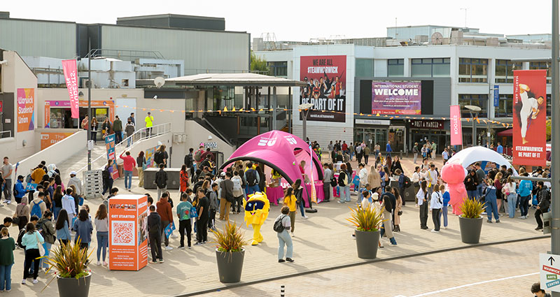 The Piazza full of people during Welcome Week, the SU tent outside and a mascot wearing a sunshine emoji